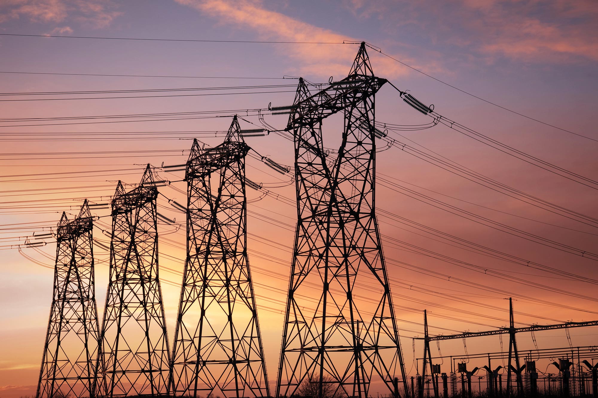High-voltage power lines and towers are silhouetted against a vibrant sunset sky with hues of orange, pink, and purple. The towers and electrical wires dominate the foreground, with a small section of utility infrastructure visible on the right.