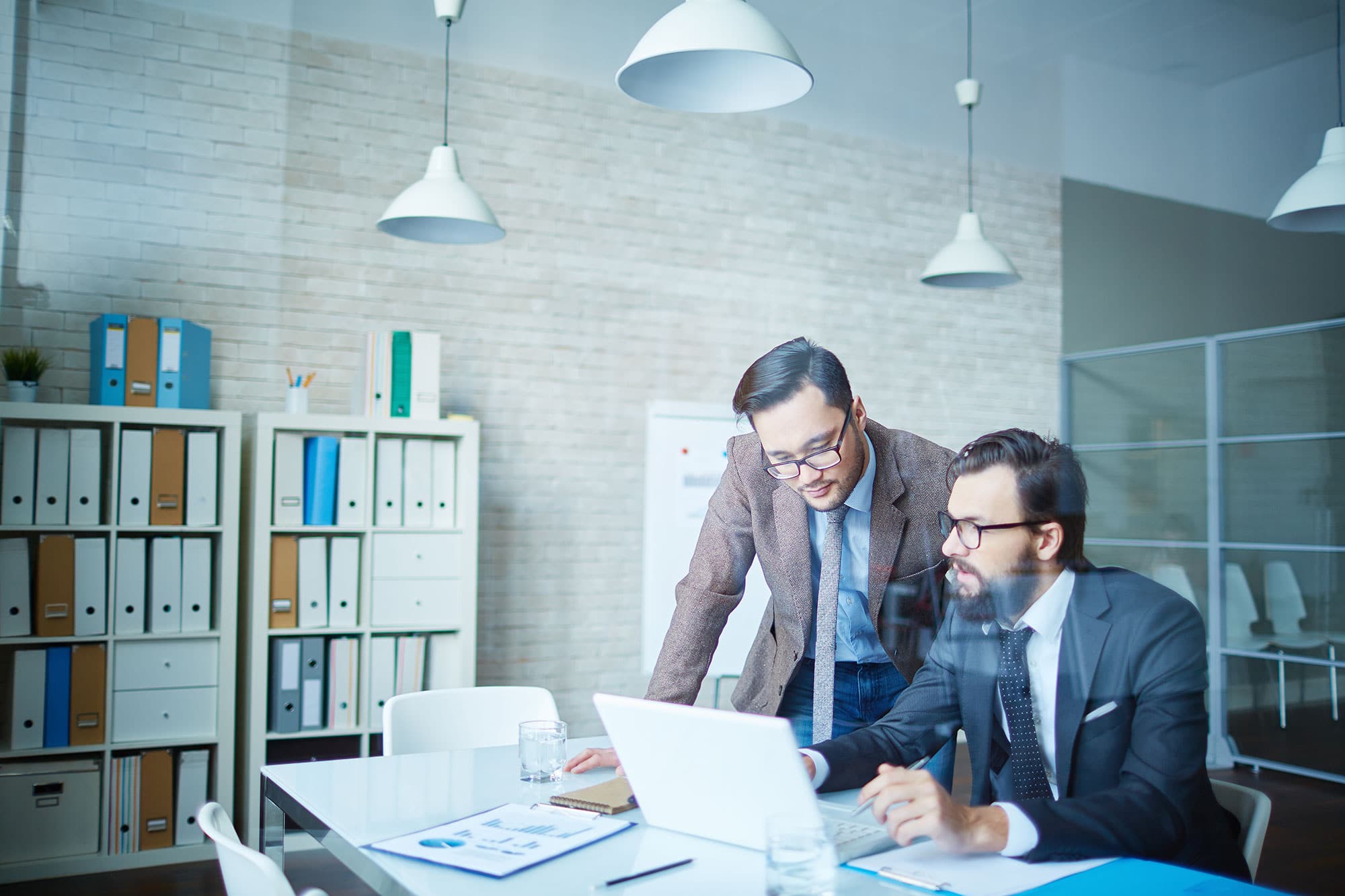 Two men in business attire are working at a modern office. One stands, leaning over to look at a laptop screen displaying "What is Gap Analysis," while the other sits and types. The office has shelves with binders, a whiteboard, large windows, and several hanging lights.