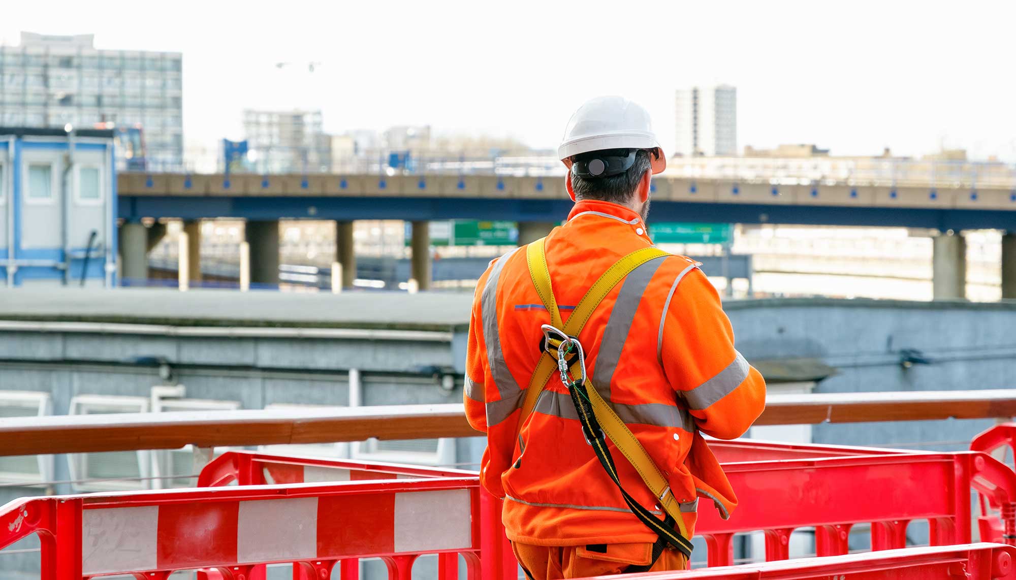 A construction worker in an orange safety jacket and white helmet stands on a raised platform beside a red safety barrier. Overlooking the UK construction site, with buildings and a bridge in the background, he witnesses the industry's growth firsthand.